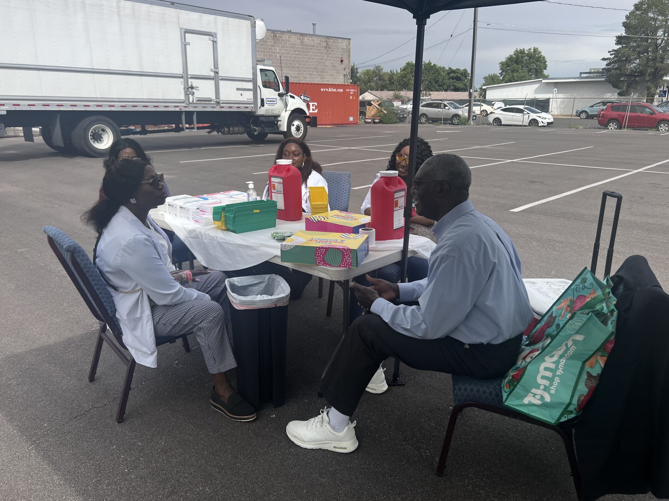 Pharmacists and volunteers seated at a supply table under a canopy at the NAPPSA flu clinic in Denver, with vaccine supplies and sharps containers on the table.